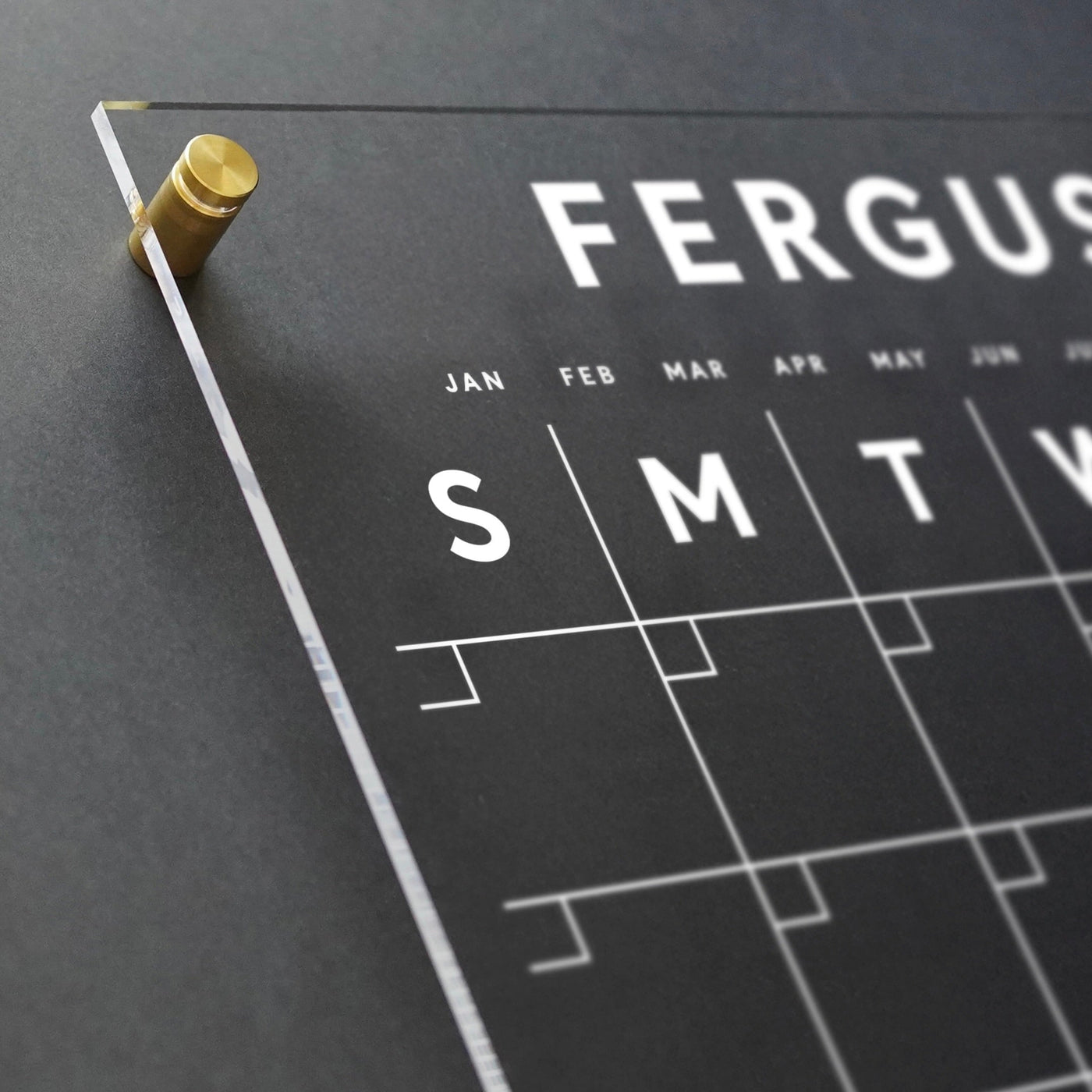 Close-up of a clear acrylic customizable monthly wall calendar by Girl Friday Shop mounted on a dark wall. It features bold white letters for months and days, secured with high-quality hardware with a gold screw; the month January is partially visible.