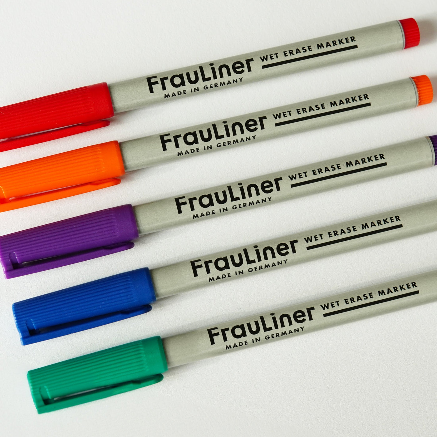 Five FrauLiner wet erase markers sit on a white surface, their caps in vivid shades of red, orange, purple, blue, and teal. With gray barrels labeled Made in Germany, theyre ideal for pairing with your sleek Girl Friday yearly dry erase acrylic calendar or a wall-mounted calendar.