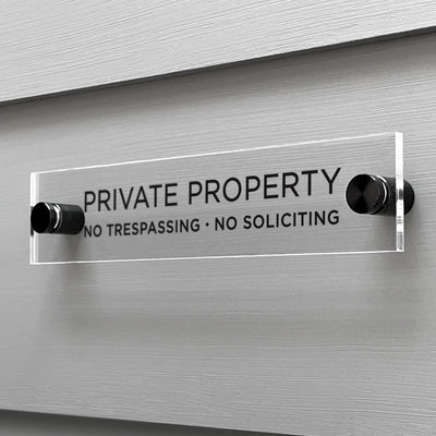 A custom Girl Friday Shop Private Property Acrylic Sign is mounted on a gray wall, stating Private Property. No Trespassing. No Soliciting in black text, and is secured with two black screws at each end.