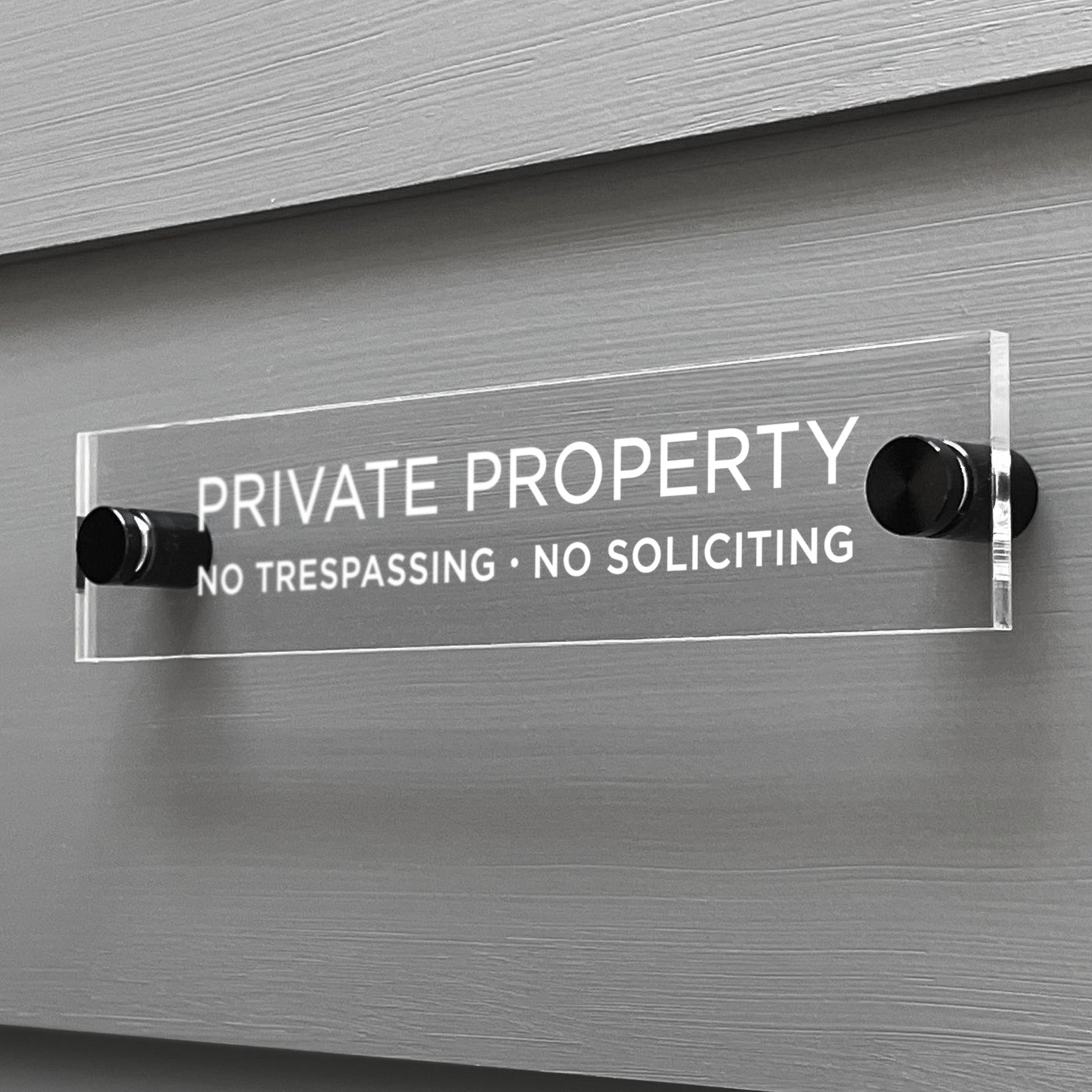 A Private Property Acrylic sign by Girl Friday Shop, featuring the text NO TRESPASSING · NO SOLICITING, is mounted on a gray wall using two black fasteners.