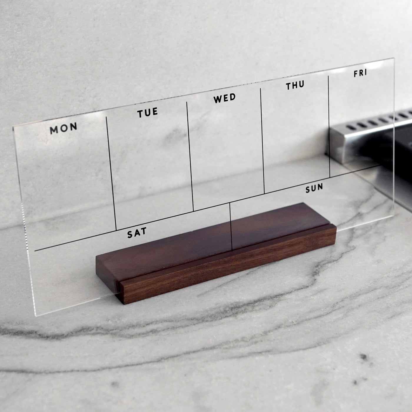 The Girl Friday Weekly Acrylic Calendar features a reusable clear acrylic planner with a wood stand, displaying sections for each day from Monday to Sunday. It elegantly rests against a marble background with days of the week printed on it.