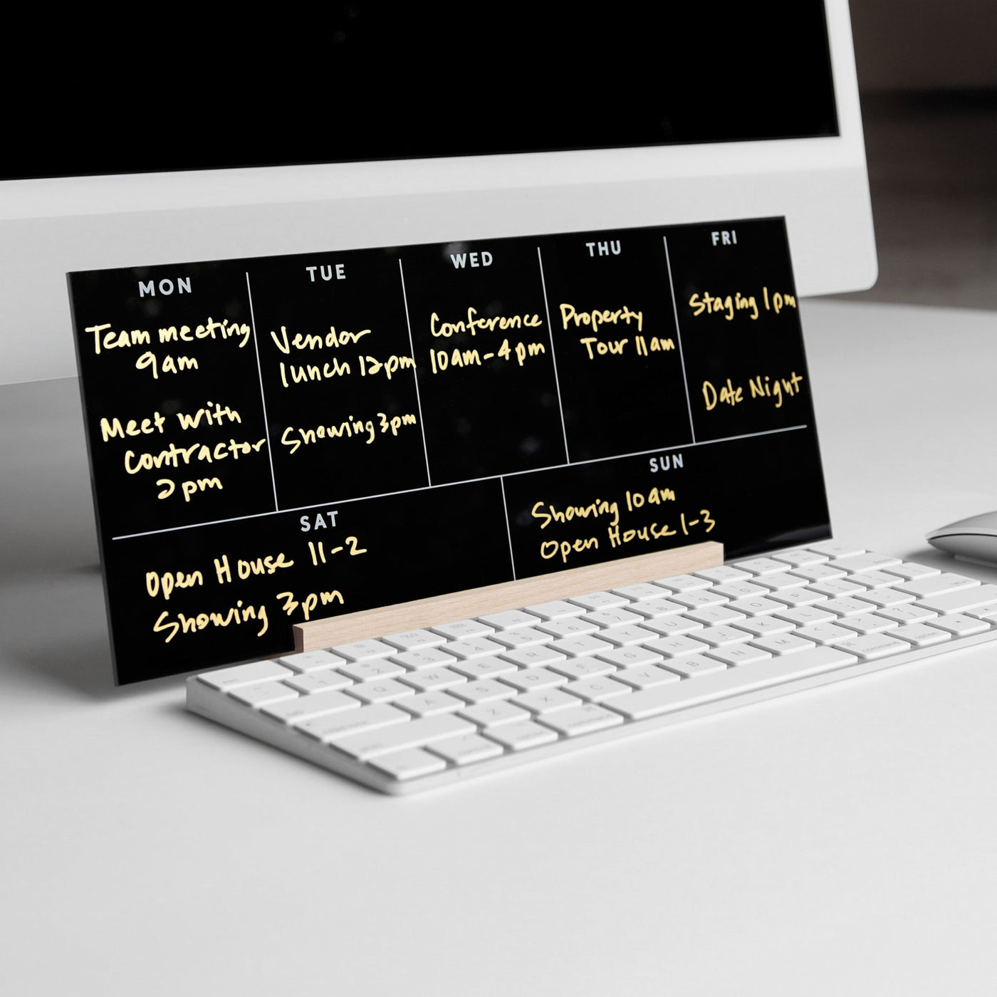 On a white desk sits the Girl Friday Weekly Acrylic Calendar with Wood Stand in sleek black, showcasing neatly inscribed gold appointments like meetings and showings. A white keyboard and monitor peek from the background.