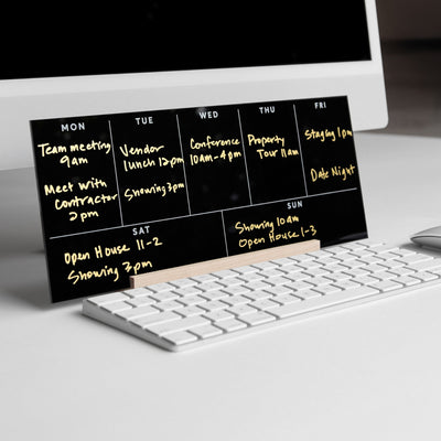 On a white desk sits the Girl Friday Weekly Acrylic Calendar with Wood Stand in sleek black, showcasing neatly inscribed gold appointments like meetings and showings. A white keyboard and monitor peek from the background.