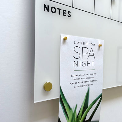 A whiteboard with NOTES at the top features a paper detailing Lilys birthday Spa Night, set for Saturday, Jan 28, 6:00 PM, at 824 Honeycut Drive. The paper is secured with stylish Gold Strong Magnets by Girl Friday Shop.