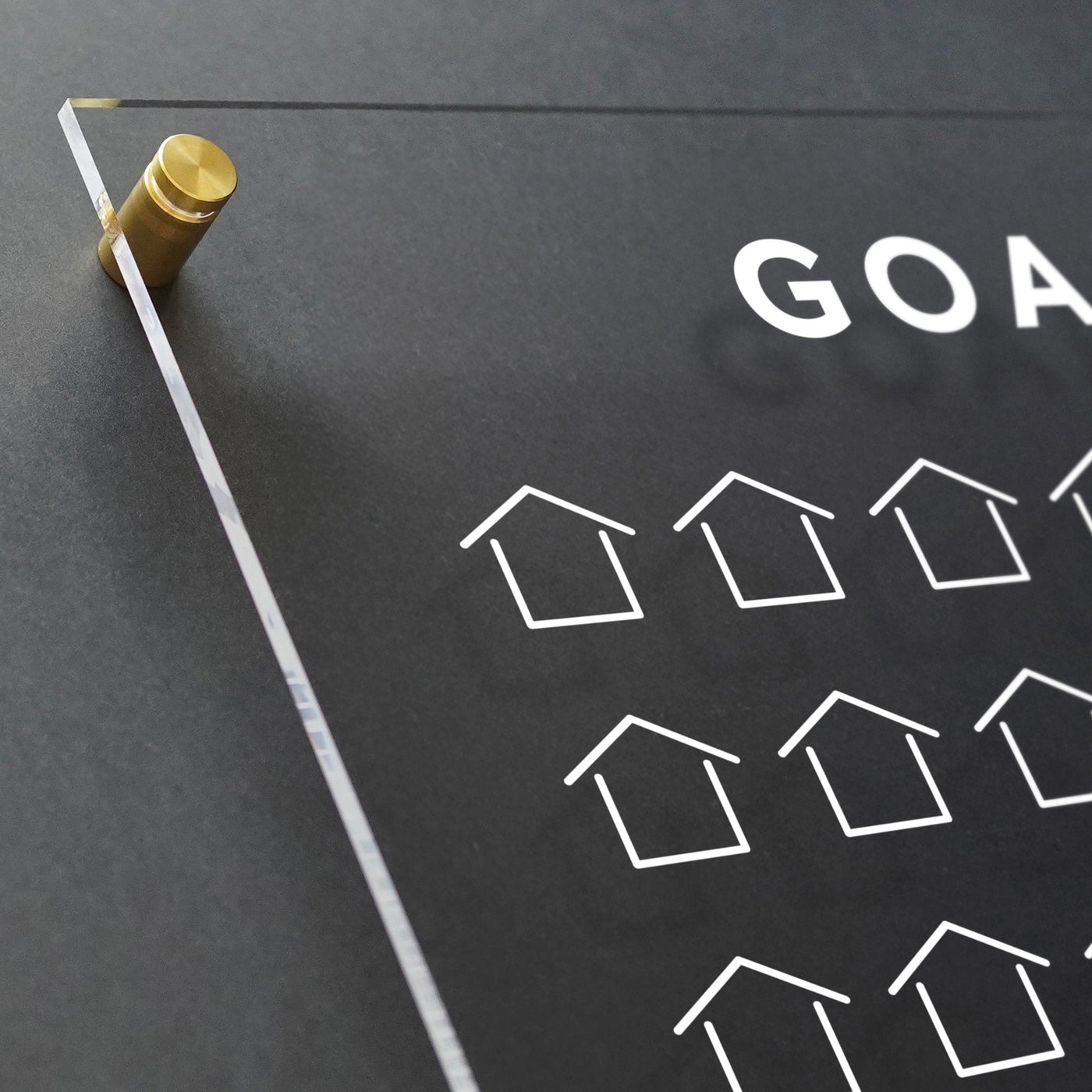The Goal Chart by Girl Friday is a clear acrylic real estate sales tracker, mounted with golden screws on a dark wall. It prominently features GOALS in white uppercase letters and includes a customizable grid of small house icons.