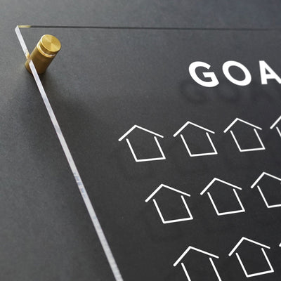 The Goal Chart by Girl Friday is a clear acrylic real estate sales tracker, mounted with golden screws on a dark wall. It prominently features GOALS in white uppercase letters and includes a customizable grid of small house icons.