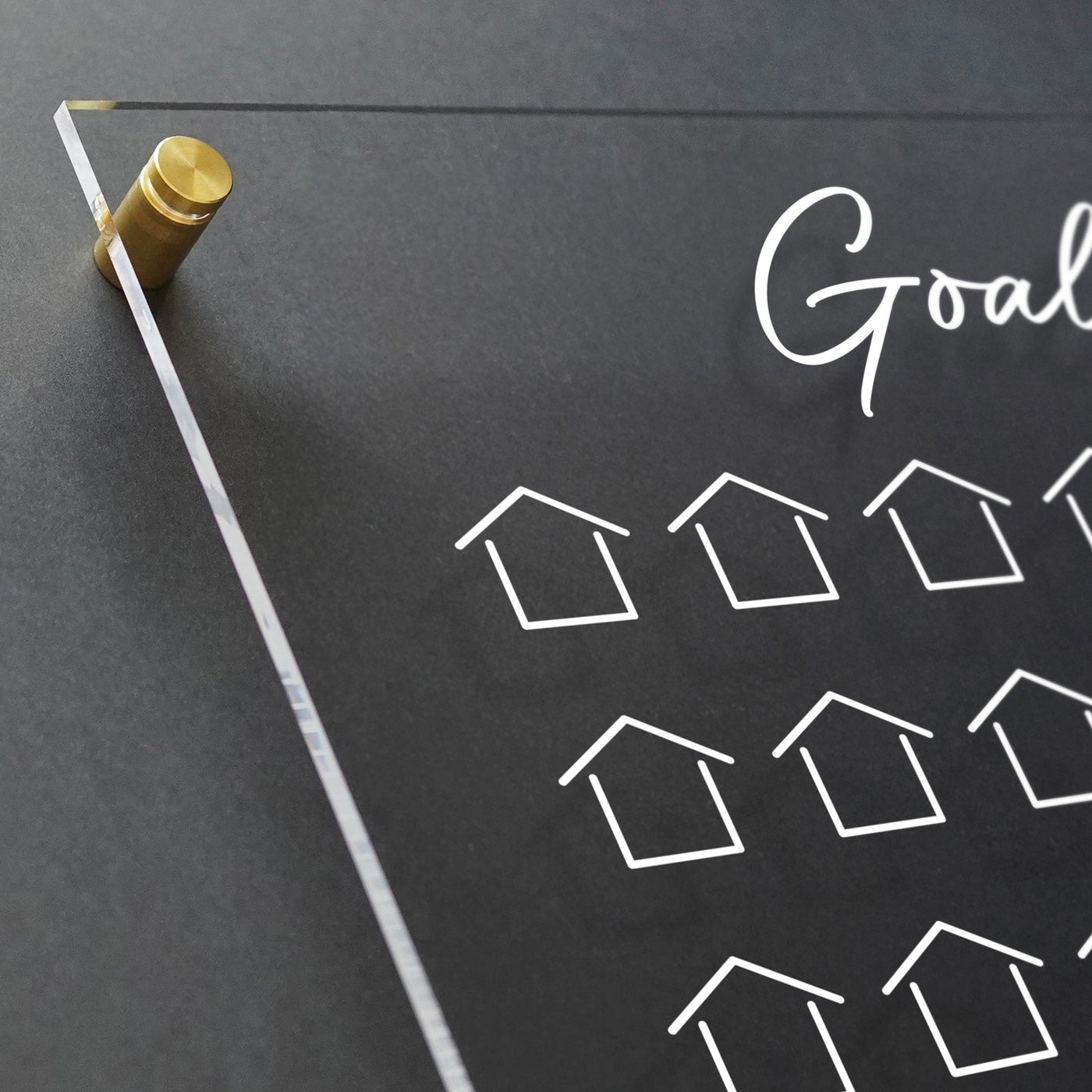 The Goal Chart by Girl Friday is a clear acrylic board with gold mounts, ideal for real estate planning. It features Goals in handwritten script and white outlined house drawings on a dark grid background, with customizable hardware for a personal touch to your plans.