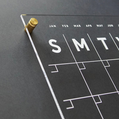 Close-up of Girl Friday Shops Acrylic Calendar with bottom notes, featuring polished edges and gold mounting hardware. The bold white day abbreviations on a black background are accompanied by month markers at the top, designed for use with wet-erase markers for convenience.