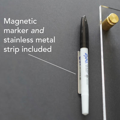 A black and white magnetic marker is set on a stainless strip against a dark gray background. Text reads, Includes magnetic marker, stainless metal strip, and wet-erase markers. On the right of the Large Acrylic Calendar by Girl Friday Shop, a gold pin accents the clear board with white text.