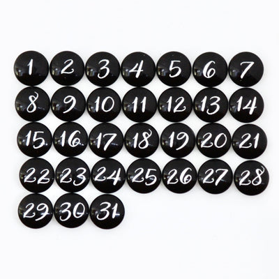 Black circular magnets numbered 1-31 in white script are arranged on a grid pattern against the premium CalendarGirlCompanys 24 x 18 Framed Whiteboard Calendar.