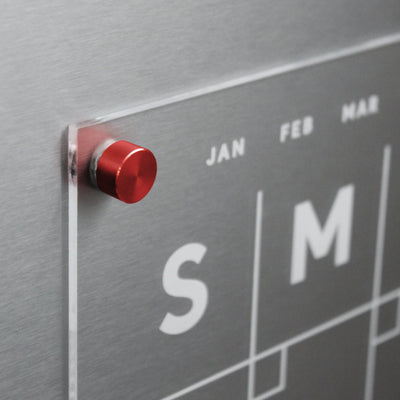 Close-up of Girl Fridays Clear Acrylic Monthly Fridge Calendar, customizable with family names and featuring white text. Beginning weeks with S and M and months like JAN, it includes a red magnetic pin for metal surfaces, perfect for notes using wet-erase markers.
