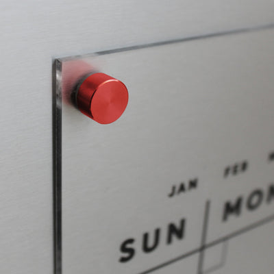 Close-up of the Girl Friday Clear Acrylic Monthly Magnetic Fridge Calendar with customizable family name. Featuring days like SUN and months like JAN, its minimalist design, held by a red magnet, pairs perfectly with wet-erase markers for easy updates.