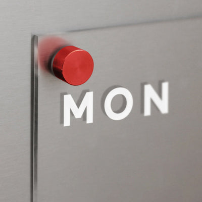 Close-up of a clear acrylic board from Girl Friday, mounted on a gray wall with MON embossed. A red circular peg secures it, casting shadows aligned with the letters. Use as an Acrylic Fridge Planner with customizable sections and dry-erase markers for daily updates.