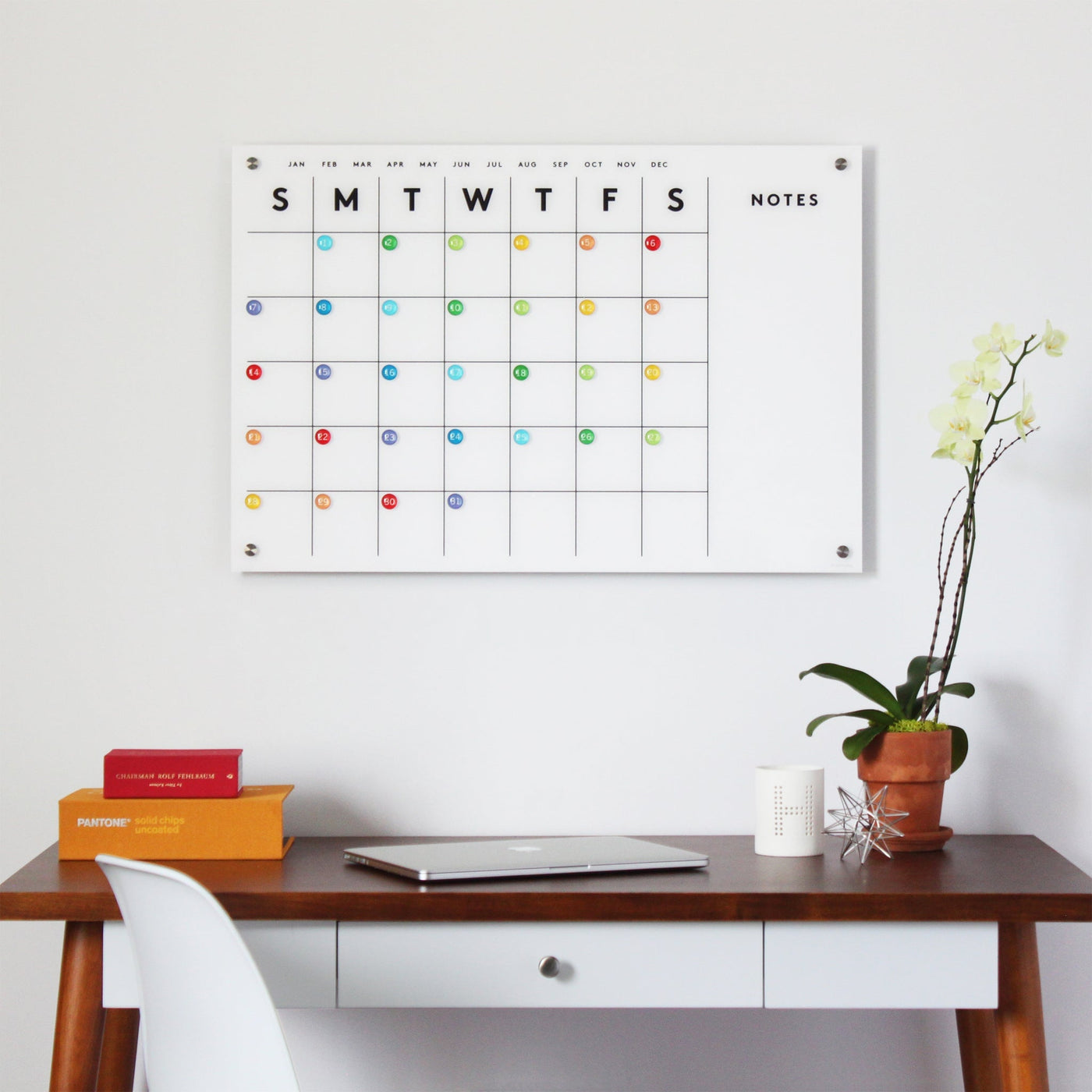 A Magnetic Acrylic Calendar with Side Notes by Girl Friday Shop hangs above the wooden desk, which features a laptop, colorful magnets, stacked books, a potted orchid, a white chair, a mug, and a small wire sculpture.