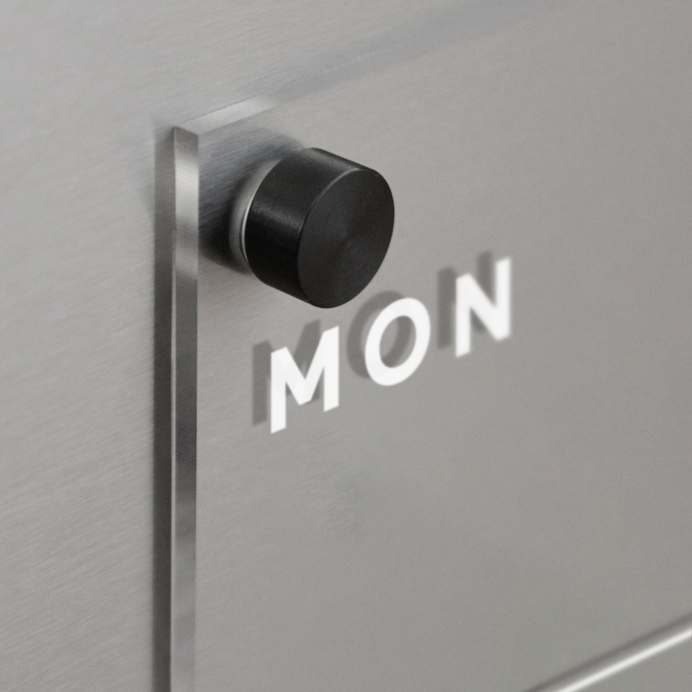 A black cylindrical knob protrudes from the matte gray surface of the Girl Friday Acrylic Fridge Planner, with MON visible for a layered effect.