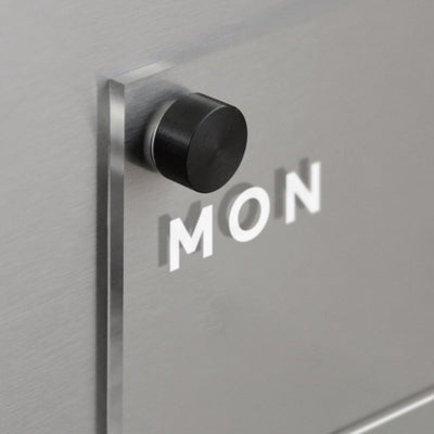 A black cylindrical knob protrudes from the matte gray surface of the Girl Friday Acrylic Fridge Planner, with MON visible for a layered effect.