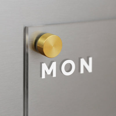 A close-up photo features a wall fixture: a clear acrylic sheet like the Acrylic Fridge Planner by Girl Friday, with MON displayed. Its mounted using a round, gold-colored metal post against a brushed metal background.