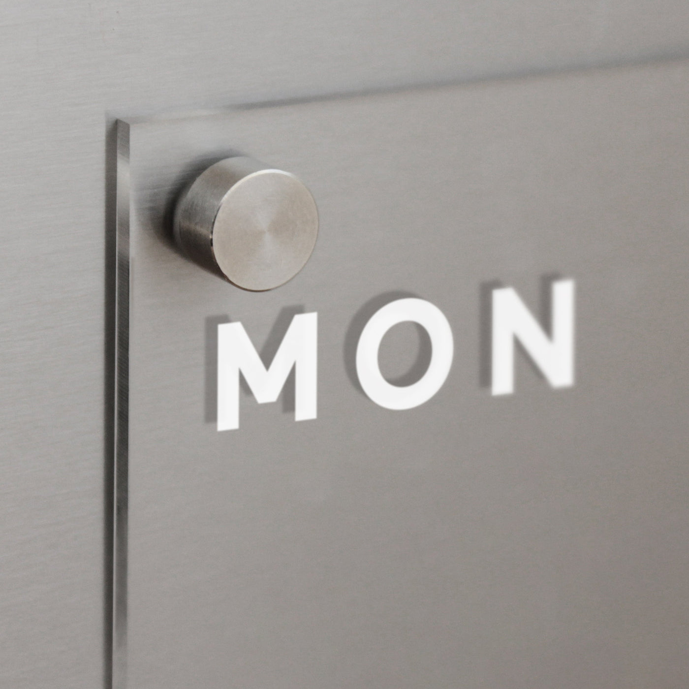 Close-up of a metallic wall with a protruding circular fixture holding Girl Fridays Acrylic Fridge Planner. The sleek panel, which casts a shadow and has MON printed on it, is customizable and pairs perfectly with dry-erase markers for easy updating.