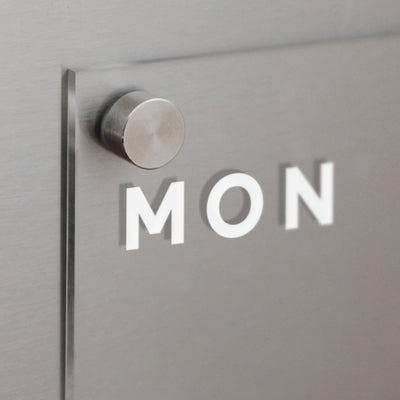 Close-up of a metallic wall with a protruding circular fixture holding Girl Fridays Acrylic Fridge Planner. The sleek panel, which casts a shadow and has MON printed on it, is customizable and pairs perfectly with dry-erase markers for easy updating.