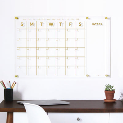 The Girl Friday Shop Clear Acrylic Monthly Wall Calendar features polished edges, gold gridlines, and a right-side NOTES section. The days are labeled S M T W T F S. Below it, a tidy desk has a closed laptop and cups of colored pencils for use with wet-erase markers.