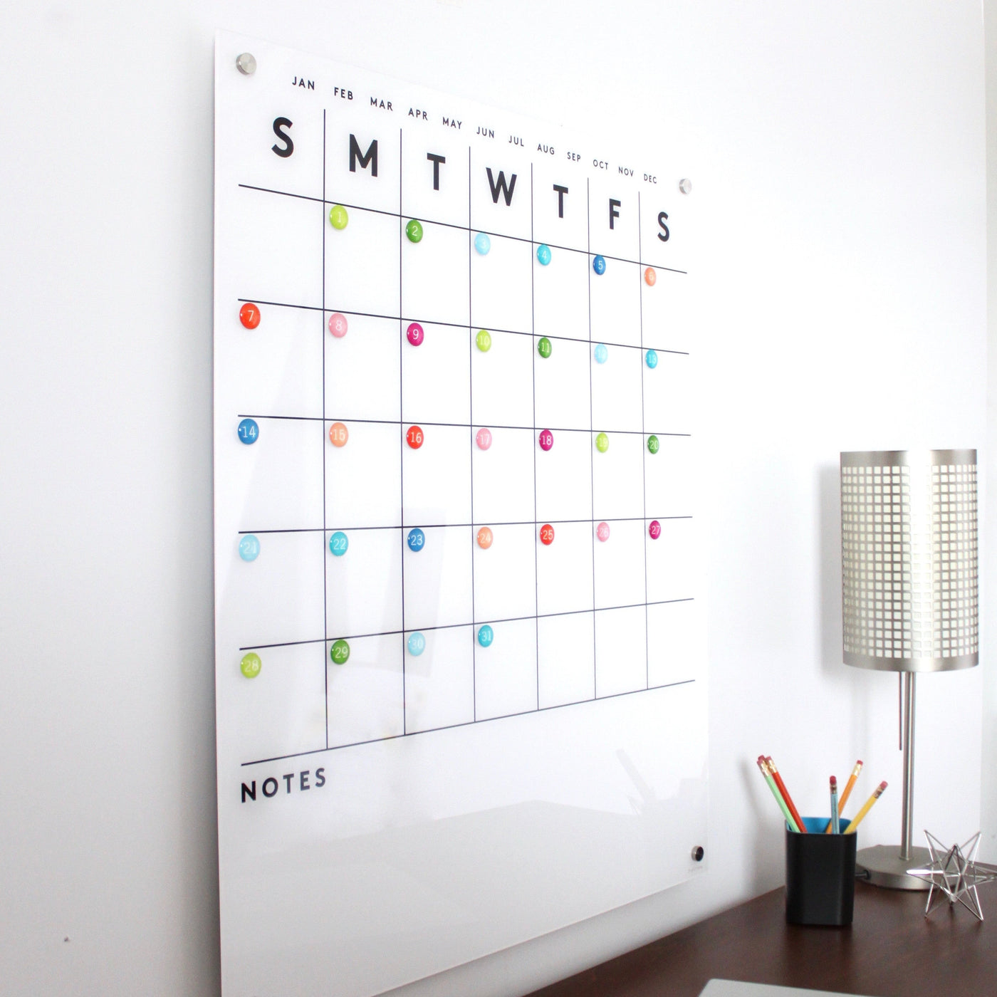 The Magnetic Acrylic Calendar with Bottom Notes by Girl Friday Shop is a wall-mounted organizer with a weekly grid. It includes colorful round magnets, labeled days, listed months above, and a notes section below. Nearby, find a lamp, cup of colored pencils, and wet-erase marker for easy updates.