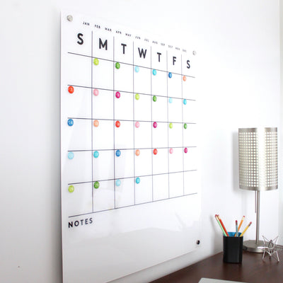 The Magnetic Acrylic Calendar with Bottom Notes by Girl Friday Shop is a wall-mounted organizer with a weekly grid. It includes colorful round magnets, labeled days, listed months above, and a notes section below. Nearby, find a lamp, cup of colored pencils, and wet-erase marker for easy updates.