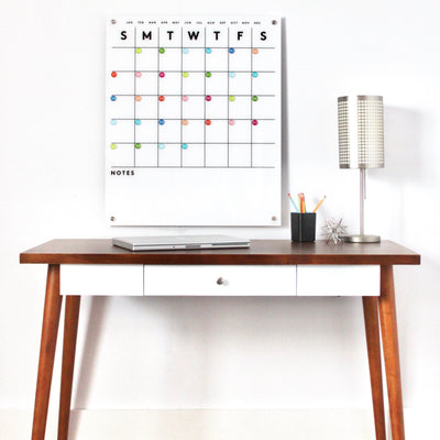 A modern wooden desk with a white drawer displays a closed laptop, a black pencil holder filled with colored pencils, and a decorative star object. Above hangs the Girl Friday Shops Magnetic Acrylic Calendar featuring colorful dots. A silver lamp is on the right.