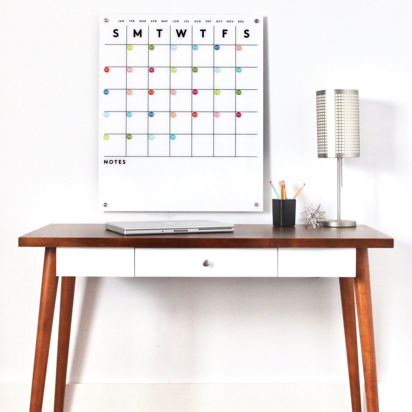 A minimalist workspace features a wooden desk with a laptop, sleek high-quality hardware, and a small black pen cup. Hanging above is Girl Friday Shops Magnetic Acrylic Calendar with colorful dots, while on the right sits a modern lamp.