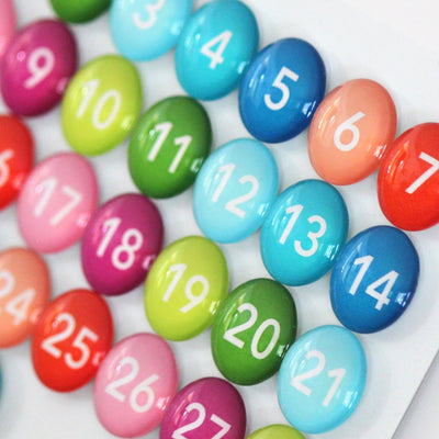 The Number Magnets by Girl Friday are colorful, round, multi-colored glass magnets numbered 1-26 featuring bold white numbers. They come in vibrant shades like reds, blues, greens, and pinks and can be arranged in a grid.