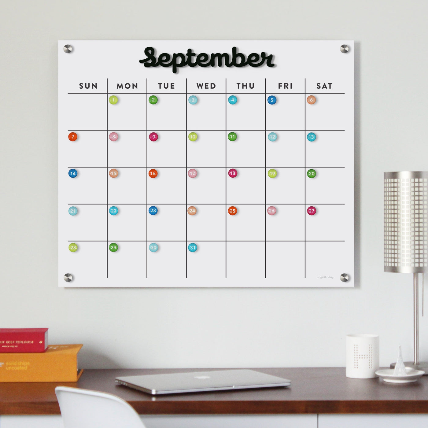 A Magnetic Acrylic Monthly Calendar by Girl Friday Shop, featuring black magnetic months, complements a tidy workspace above a minimalist desk. Paired with a modern metallic lamp and books, it offers an organized planning surface for September.