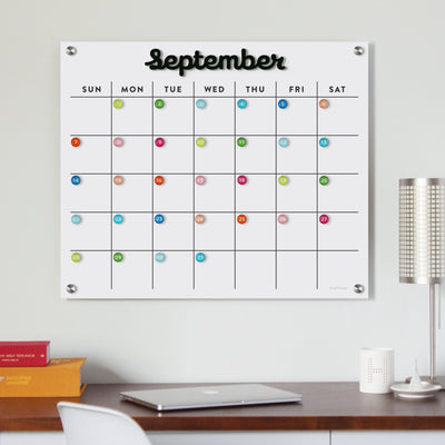 A Magnetic Acrylic Monthly Calendar by Girl Friday Shop, featuring black magnetic months, complements a tidy workspace above a minimalist desk. Paired with a modern metallic lamp and books, it offers an organized planning surface for September.