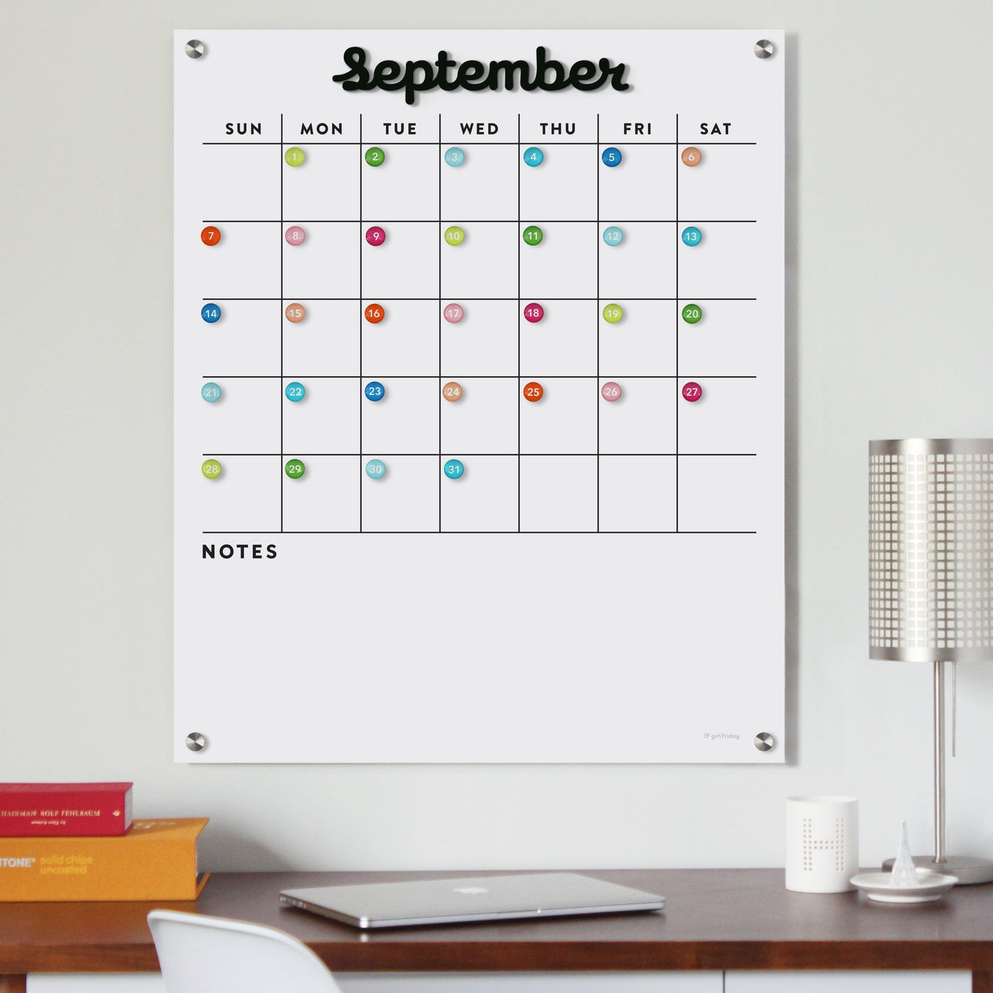 The Girl Friday Shops Magnetic Acrylic Calendar features a grid layout with black magnetic months and colorful pushpins for each day, plus blank Notes sections. Its complemented by a modern lamp and a closed red book, creating an organized desk setup for September.