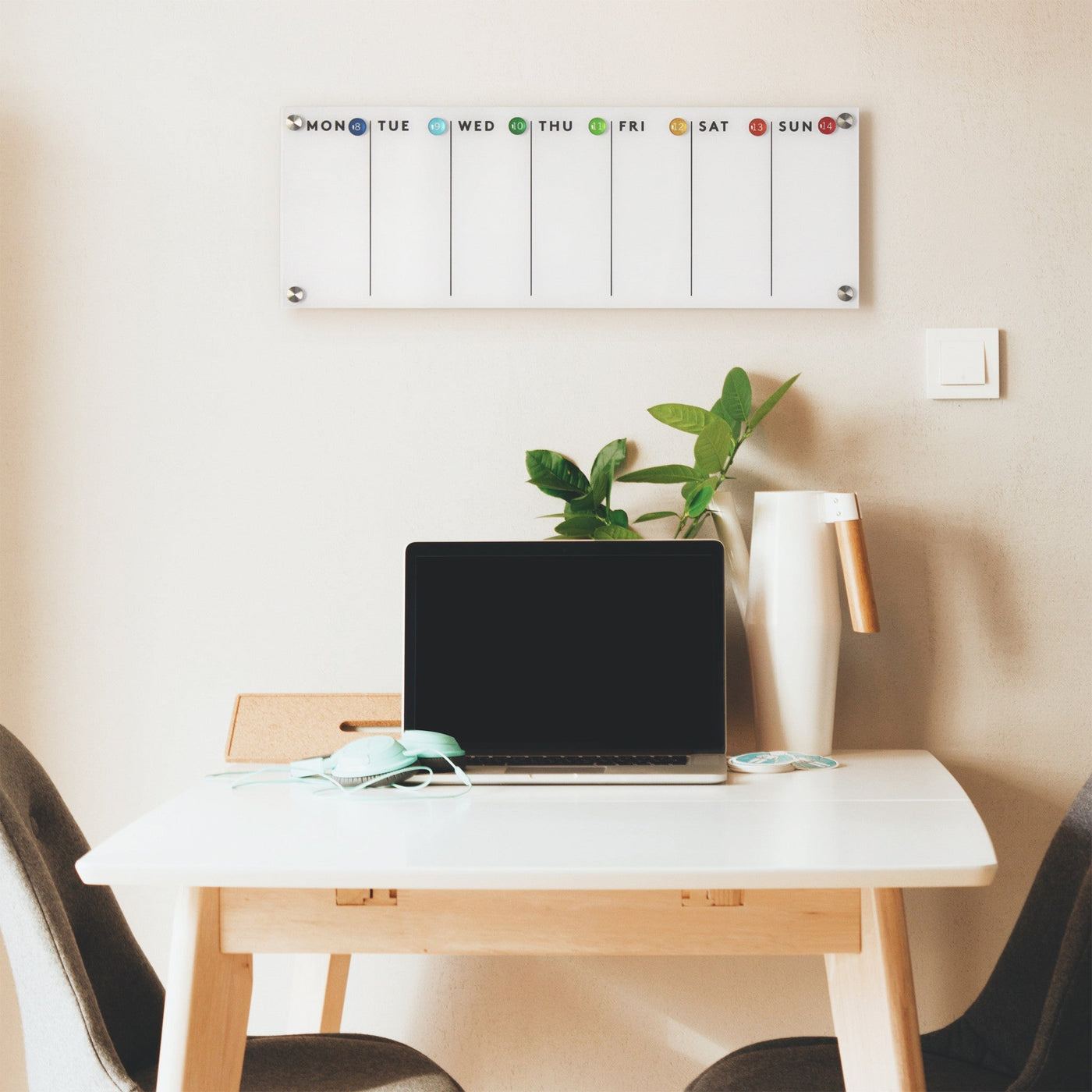 A minimalist workspace features a wooden desk with a laptop, headphones, and a plant in a vase. Above hangs the Girl Friday Shops sleek Magnetic Acrylic Calendar | Weekly for planning ease. Two grey chairs sit by the beige wall, near a visible light switch.