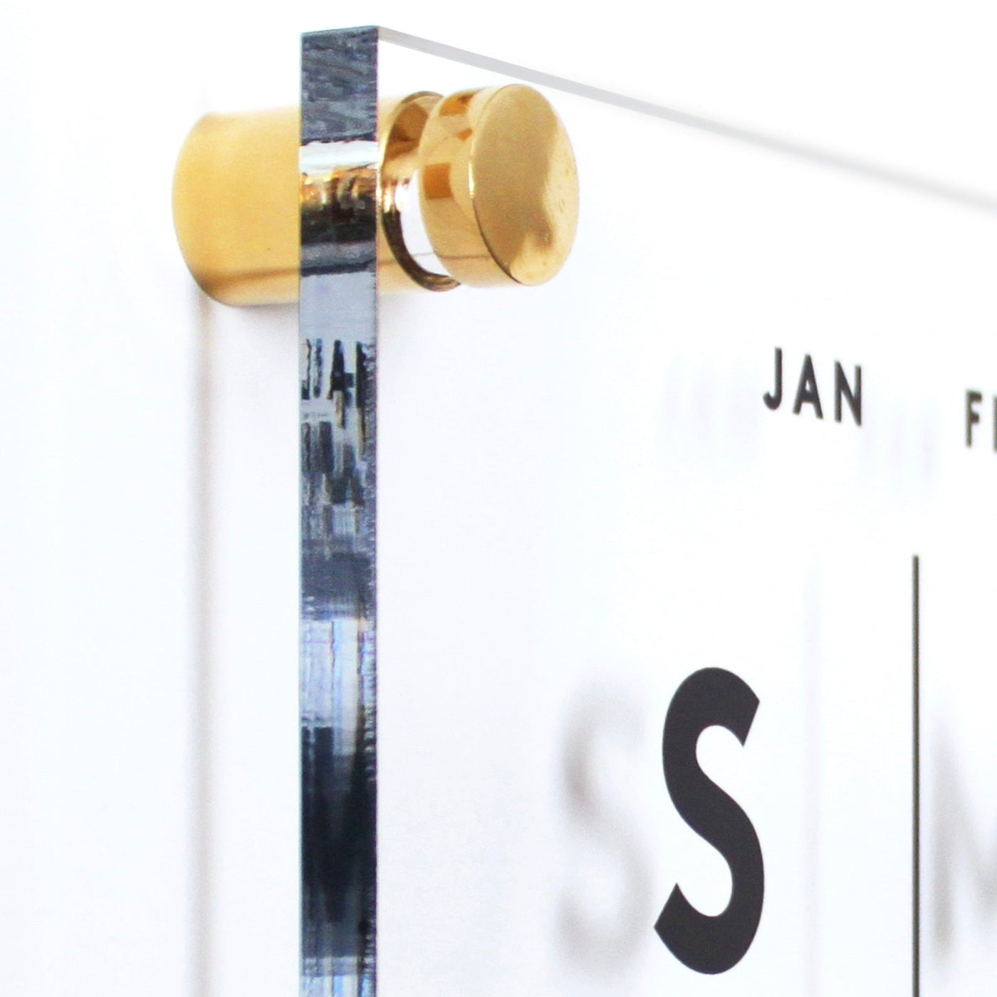 A close-up of the Girl Friday Clear Acrylic Monthly Wall Calendar shows it mounted with high-quality hardware and a gold bracket. S and partial months JAN and FEB are visible, with a blurred edge reflection—a perfect surface for wet-erase markers.