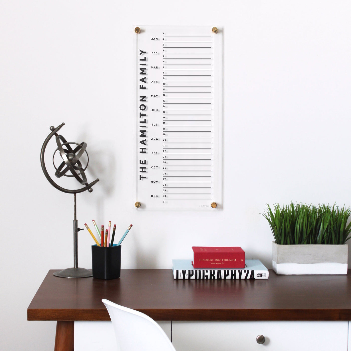 A desk features a globe figurine, a cup with colored pencils, and three books including TYPOGRAPHY 2. Above is Girl Friday Shops Clear Acrylic personalized monthly calendar highlighting family birthdays. A potted plant on the right adds greenery.
