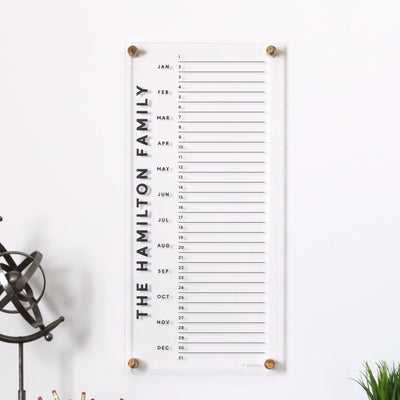 The Clear Acrylic personalized monthly calendar by Girl Friday Shop displays The Hamilton Family on the left, crafted from high-quality materials. It lists months from January to December, with space for dates, and a small sculpture adds elegance.