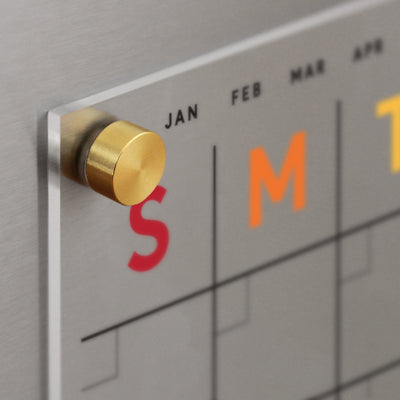 Close-up of the Clear Acrylic Monthly Fridge Calendar by Girl Friday, showing a transparent panel with a brass knob. Abbreviations for January, February, and March are visible at the top, while the days S and M appear in red and orange rainbow text.