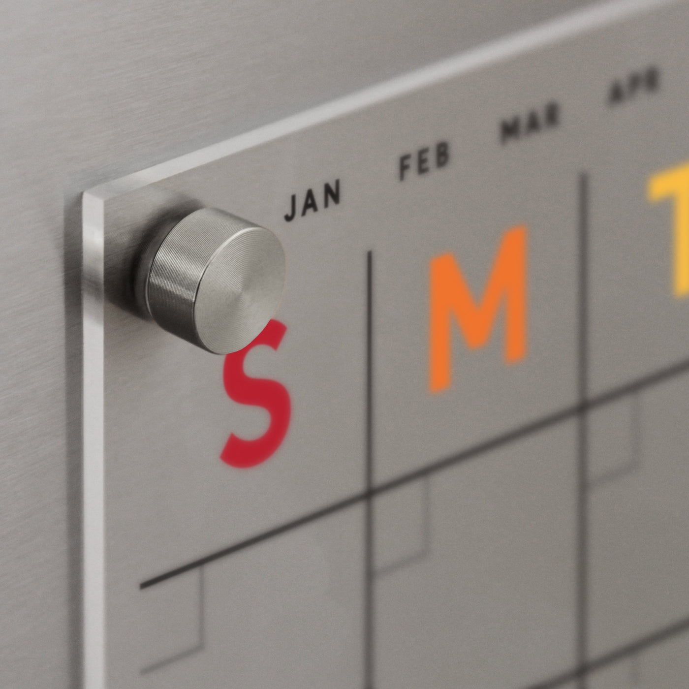 Close-up of the Clear Acrylic Monthly Fridge Calendar by Girl Friday, featuring metallic knobs and stainless steel magnets. S and M are highlighted in red and orange on the sleek, minimalistic design. Perfect for wet-erase markers, its an ideal addition to any modern kitchen space.