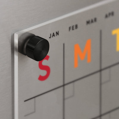 Close-up of a Clear Acrylic Monthly Fridge Calendar | Rainbow Text by Girl Friday, highlighting the top left corner with JAN, FEB, MAR, and APR. Secured with sleek stainless steel magnets, the red S and orange M stand out.