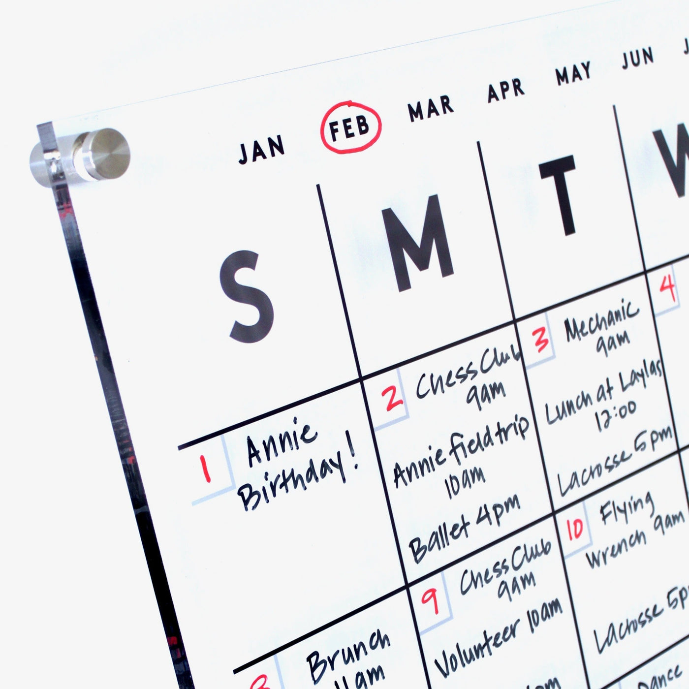 The Girl Friday Shops Clear Acrylic Monthly Wall Calendar elegantly displays February, featuring handwritten notes like Annie Birthday! in red on the 1st and reminders for Chess Club, Ballet, Mechanic 9am, and Volunteer 10am. Sturdy screws securely hold this quality fixture.
