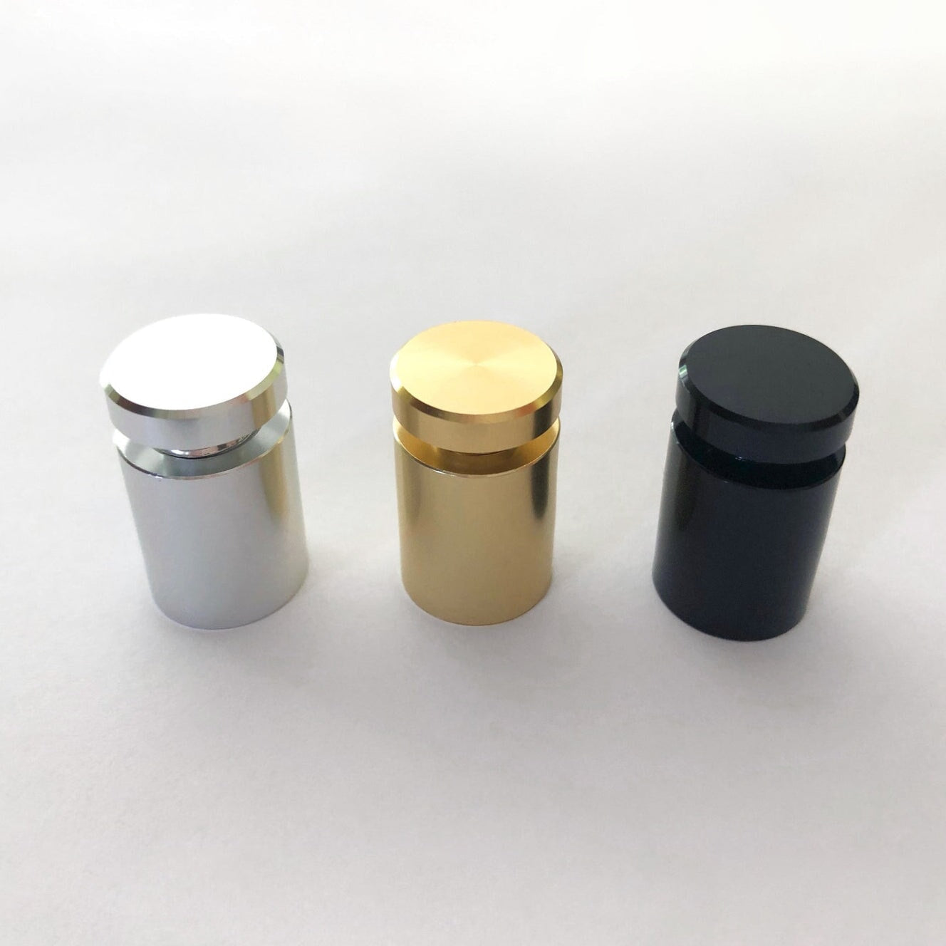Three cylindrical standoffs, crafted from high-quality materials, are displayed on a white surface. From left to right, they are silver, gold, and black, boasting smooth reflective surfaces and flush tops. Perfect for enhancing home organization with style and elegance using the Clear Acrylic Monthly Wall Calendar with Notes from Girl Friday Shop—now 60% off!.