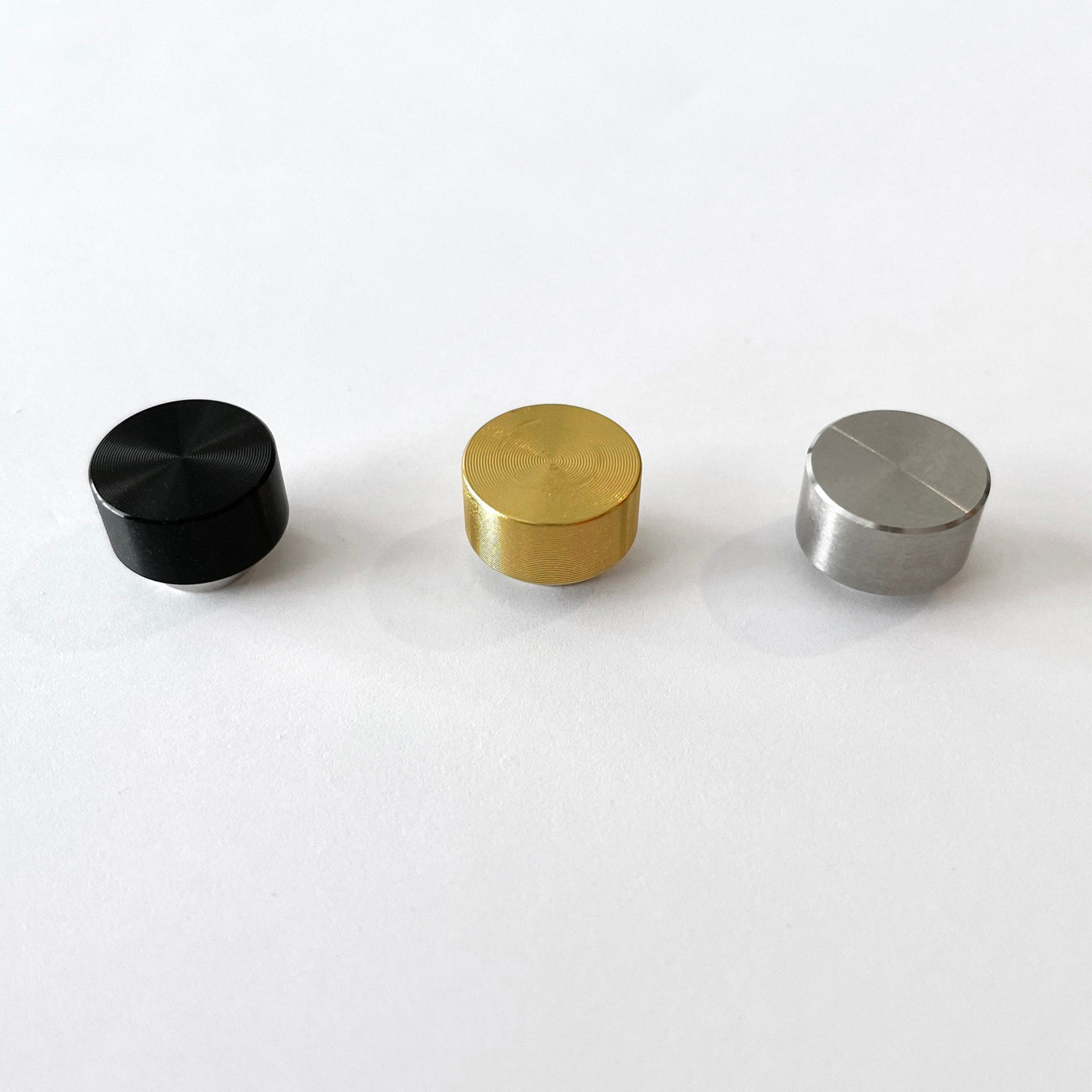 Three small cylindrical knobs in black, gold, and silver are lined up on a white surface from left to right. Each has a slightly reflective finish reminiscent of the gleaming accents in the Girl Friday Acrylic Fridge Calendar weekly.