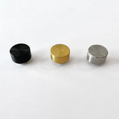 Three small cylindrical knobs in black, gold, and silver are lined up on a white surface from left to right. Each has a slightly reflective finish reminiscent of the gleaming accents in the Girl Friday Acrylic Fridge Calendar weekly.